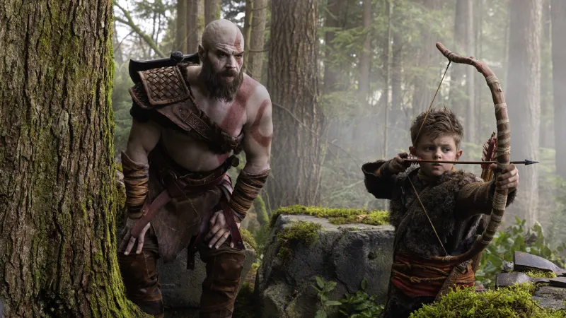 Kratos watches his son Atreus aim a bow. Kratos has a sort of "baseball coach dad" stance, leaning forward with his hands on his knees.